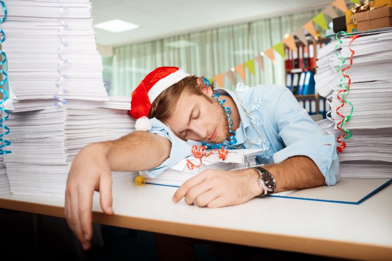 Tired young handsome businessman in red santa claus hat sleeping at workplace among papers on christmas day.